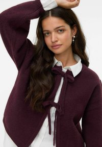 Cardigan en maille bordeaux avec une coupe décontractée, doté d'une fermeture à nouer, de manches longues et d'une texture douce superposée à une chemise blanche.