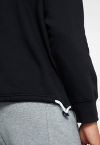 Felpa nera con texture liscia, caratterizzata da coulisse all'orlo e una vestibilità aderente. Abbinata a pantaloni grigio chiaro.