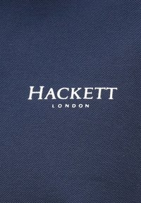 Jūras spēku audums ar teksturētu virsmu, ar baltu izšūtu tekstu "HACKETT" virs "LONDON."