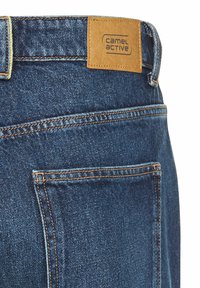 Denim jeans med en medium vask, der har et tan lædermærke med teksten "camel active" og kontrastsyninger langs sømmene og lommerne.