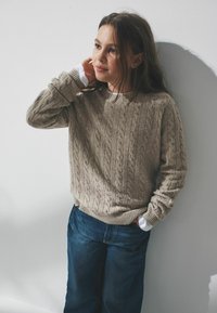 Jasno beżowy sweter o splocie warkoczowym z prążkowanymi mankietami i dekoltem, w połączeniu z luźnymi niebieskimi dżinsami. Teksturowany materiał ukazuje skomplikowane wzory.