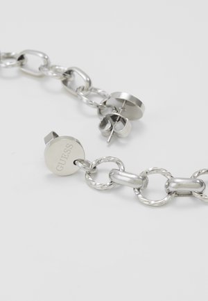 Zilveren ketting met gestructureerde en gladde ovale schakels en een rond Guess-bedeltje eraan bevestigd, liggend op een wit oppervlak.