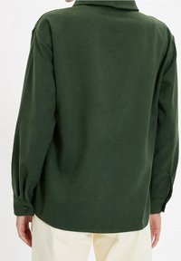 Camicia verde a maniche lunghe in un tessuto liscio e leggero, con colletto classico e polsini abbottonati. L'orlo è leggermente arrotondato.