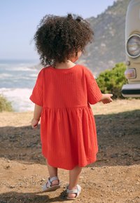 Kind in einem roten, strukturierten Baumwollkleid mit kurzen Ärmeln, geraffter Taille und grauen Sandalen, mit dem Rücken zu einem Strand und einem Vintage-Fahrzeug gewandt.