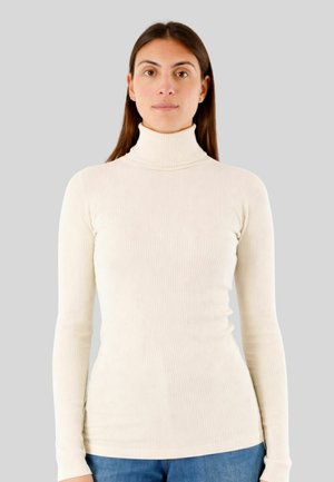 Donna con capelli castano lisci che indossa un dolcevita a coste color crema e jeans blu, in piedi davanti a uno sfondo grigio neutro.