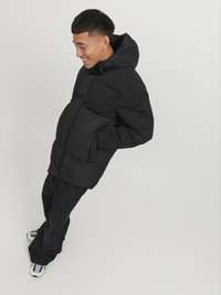 Zwarte pufferjack met capuchon, met een gewatteerd ontwerp en voorzakken. Model draagt een bijpassende donkere broek en gestreepte sneakers.