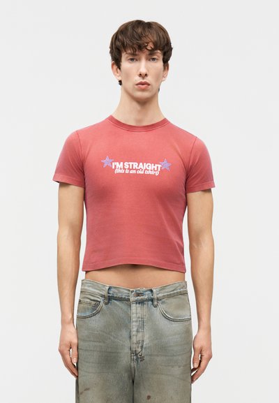 Mladý muž nosiaci krátke červené tričko s bielym nápisom „I'M STRAIGHT (toto je staré tričko)“ a vyblednuté modré rifle, stojaci pred jednoduchým pozadím.