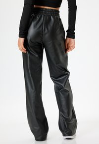 Pantaloni neri in ecopelle con un'ampia fascia elastica in vita, gamba larga e una superficie liscia e lucida, mostrati da dietro.