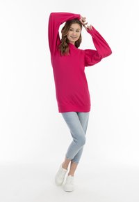 Top lungo rosa brillante con maniche a sbuffo e colletto alto, abbinato a jeans skinny azzurri chiari e sneakers bianche. Texture liscia.