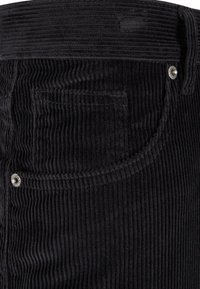 Gros plan sur un tissu en velours côtelé noir avec une poche visible, des surpiqûres et des rivets métalliques sur un pantalon ou une jupe.