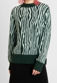 Grüner Pullover mit einem strukturierten Muster, das Holzmaserung ähnelt, mit hellen Streifen und gerippten Bündchen und Saum. Gemütliches Strickmaterial.