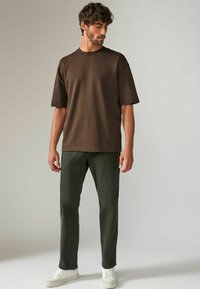 T-shirt en coton marron à manches courtes, coupe décontractée, associé à un pantalon vert foncé et des baskets blanches. Design simple et épuré.