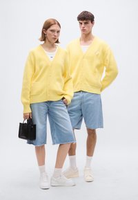 Κίτρινες ριπ πλεκτές ζακέτες με κουμπιά, σε συνδυασμό με ανοιχτόχρωμα τζιν σορτς. Το μοντέλο κρατάει μια μαύρη τσάντα χειρός, με λευκά αθλητικά παπούτσια και κάλτσες.