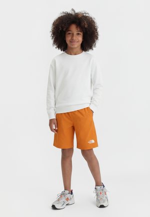 Enfant aux cheveux bouclés portant une chemise blanche à manches longues, un short orange et des baskets blanches, debout avec une main dans la poche.