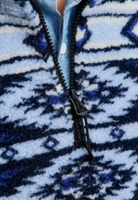 Polarowy sweter w jasnoniebieskim kolorze z granatowym i białym abstrakcyjnym wzorem, posiada czarny zamek błyskawiczny z uchwytem oraz dżinsową koszulę pod spodem.
