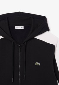 Sweat-shirt à capuche noir avec fermeture éclair, panneaux blancs sur les épaules, cordons de serrage, et petit logo crocodile vert sur la poitrine.