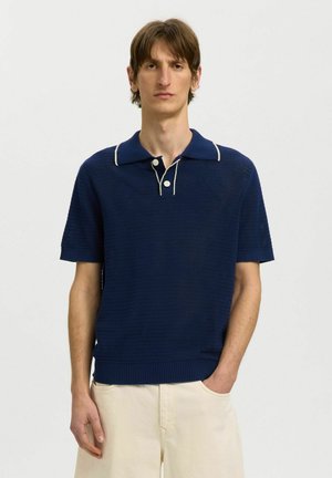 Homme à la peau claire et aux cheveux bruns portant un polo en maille bleu marine à manches courtes et un pantalon beige sur fond blanc uni.