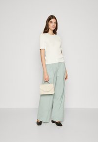 λευκό υφασμάτινο κοντομάνικο μπλουζάκι συνδυασμένο με μίντι πράσινα φαρδιά παντελόνια. Κρατάει μια μπεζ τσάντα χειρός με διακριτό σχέδιο φάκελου. Μαύρα παπούτσια.