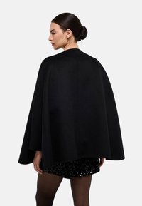 Cape en laine noire avec un col montant et une coupe fluide, s'étendant jusqu'au milieu des cuisses. Texture lisse et lignes épurées ; garniture minimale.