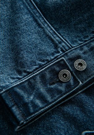 Tumma sininen farkkukangas, jossa on hieman haalistunut rakenne. Varustettu hopeanvärisillä metallinapeilla, joissa lukee "OMBRE" ja "DENIM." Lähikuva hihansuusta.