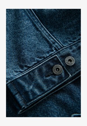 Tumma sininen farkkukangas, jossa on hieman haalistunut rakenne. Varustettu hopeanvärisillä metallinapeilla, joissa lukee "OMBRE" ja "DENIM." Lähikuva hihansuusta.