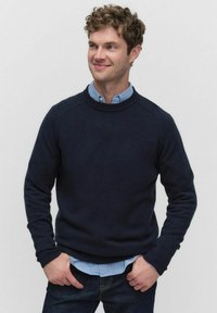 Marinblå stickad tröja med ribbad halsringning och muddar, bärs över en ljusblå skjorta med krage, kombinerad med mörka jeans. Slät textur.