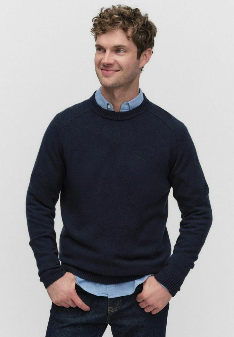 Marinblå stickad tröja med ribbad halsringning och muddar, bärs över en ljusblå skjorta med krage, kombinerad med mörka jeans. Slät textur.