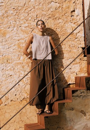 Femeie cu tatuaje, purtând pantaloni largi maro și top fără mâneci în carouri, stă pe o scară metalică rustică, în fața unui perete texturat de piatră.