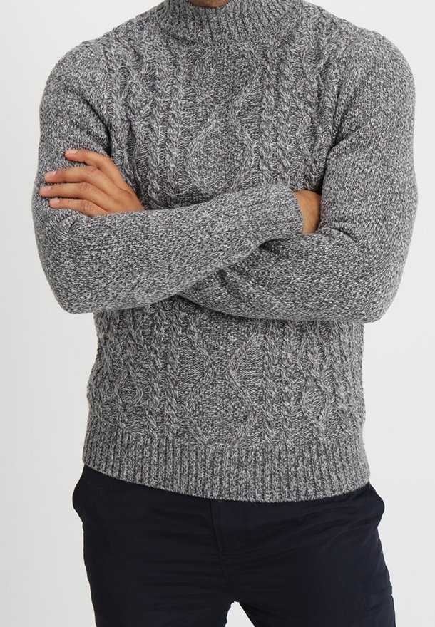 Homme aux bras croisés portant un pull gris à torsades et un pantalon sombre, debout devant un fond clair uni.