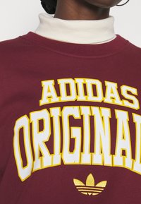 Vinröd sweatshirt med vit och gul "ADIDAS ORIGINAL" text. Har rund halsringning och mjuk textur. Inkluderar Adidas treflöj-symbol.