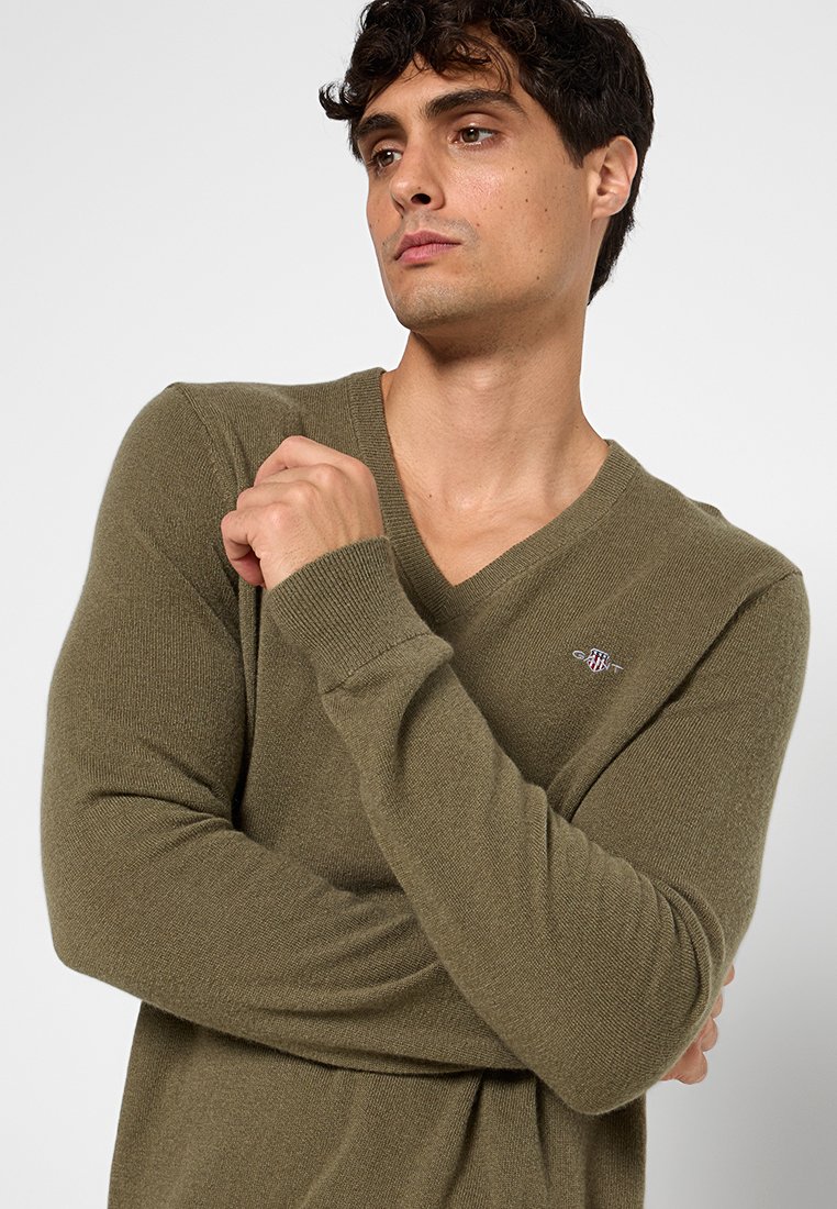 Pull à col en V vert olive en tissu doux, présentant des manches longues et un petit logo sur la poitrine. Design en tricot texturé.