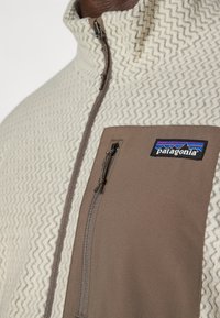 Fleece bunda s krémovým vzorom v zigu-zagu a hnedým panelom, má zipsovú kapsu a obsahuje logo Patagonia na hrudi.