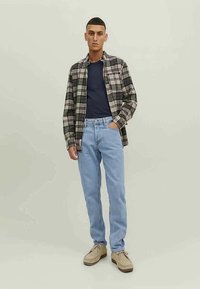 Rutig flanellskjorta i svart, grå och krämfärgad över en marinblå T-shirt; ljusblå jeans; beige skor. Modellen står mot en neutral bakgrund.