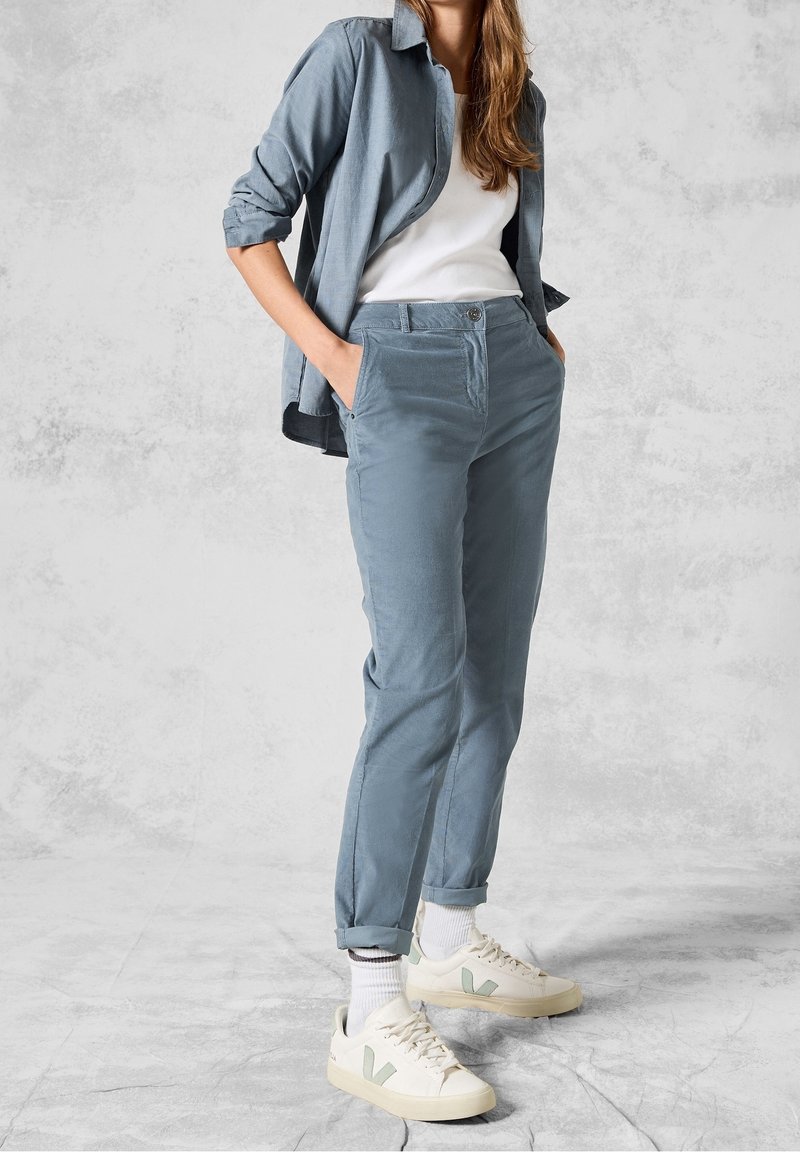 Lichtblauwe katoenen broeken met zijzakken, gecombineerd met een lichtblauw shirt en een wit T-shirt. Schoenen zijn beige met groene accenten.