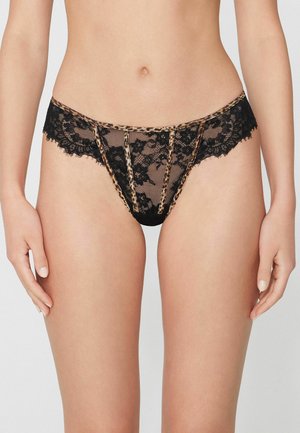 Donna che indossa biancheria intima nera in pizzo con rifiniture leopardate, mostrata dalla vita a metà coscia, in piedi davanti a uno sfondo semplice.