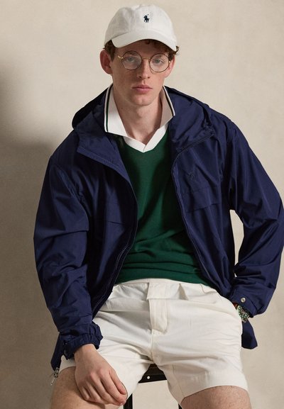 Jeune homme portant des lunettes rondes, une casquette blanche, une veste bleu marine, un pull vert par-dessus une chemise blanche à col et un short blanc, assis devant un fond uni.