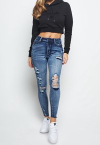 Mujer vestida con un sudadera corta negra, jeans ajustados azules desgastados y zapatillas blancas, de pie contra un fondo blanco liso.