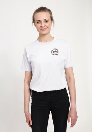 Smilende kvinde med lyst hår i en hvid t-shirt med "Liquid Engineering Castrol"-logo og sorte bukser mod en ens hvid baggrund.