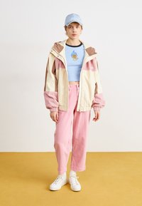 Farverig kordjakke i creme, pink og brun, parret med en lyseblå crop top med flammedesign og pink bukser. Hvite sneakers fuldender looket.