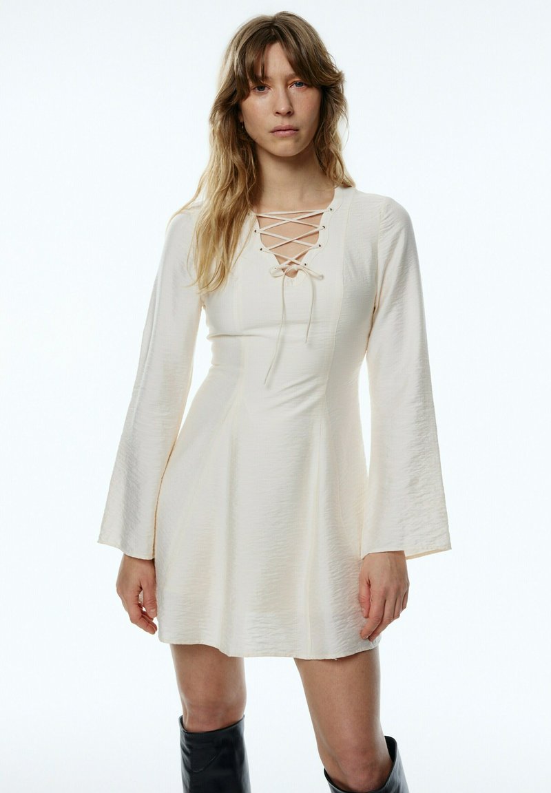 Cremefarbenes Kleid mit Schnürung, langen, ausgestellten Ärmeln, strukturiertem Stoff und tailliertem Oberteil, das eine A-Linienform schafft und oberhalb des Oberschenkels endet.