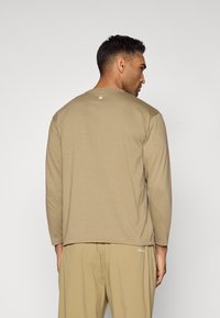 Long-sleeve beige shirt made of soft fabric with a crew neck. Features a small white logo above the neck and relaxed fit.