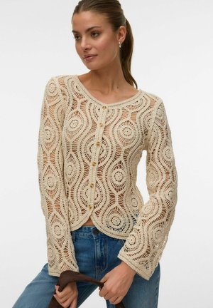 Femme portant un haut beige en crochet à manches longues et boutonné, associé à un jean bleu, tenant un objet brun devant un fond uni.