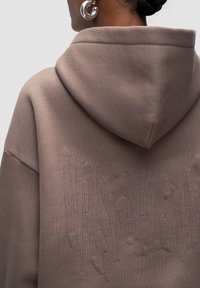 Oversized hoodie in taupe, featuring a hood and textured stitching of abstract shapes across the back. Smooth fabric with a comfy fit.