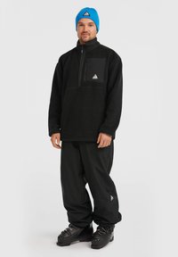 Pullover en polaire noir avec une poche poitrine zippée, associé à un pantalon noir ample et des boots de ski noires. Bonnet bleu avec un détail logo triangulaire.