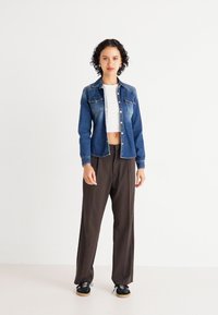 Giacca in denim blu con chiusura a bottoni, abbinata a una camicia bianca cropped e pantaloni marroni larghi. Sneakers nere completano il look.