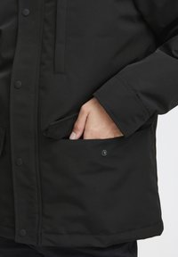 Sort polstret jakke med et stilrent design, der har en sidelomme og trykknaplukninger. Stoffet har en glat, mat tekstur.