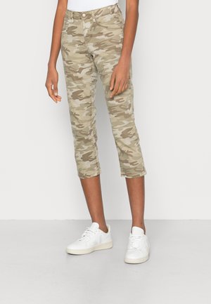 Calças cropped com padrão camuflado em tons de bege e verde, apresentando um corte slim, fecho de zip e cinco bolsos. Combinadas com ténis brancos.