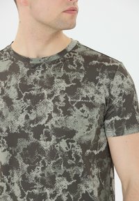 Grå och grön camouflage-mönstrad sporttröja, ribbad textur, korta ärmar, rund halsringning, har "ENDURANCE"-logotypen på bröstet.