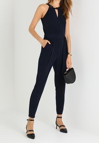 Marinblå overall med halterneck och nyckelhålsdetalj, med en midja som är insvängd, avsmalnande ben och två sidofickor. Matchad med svarta klackar och en handväska.