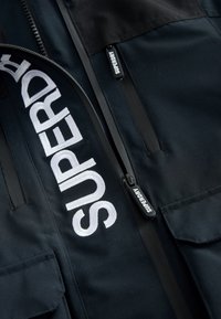 Must mustriga väliskate, millel on suur valge tikandiga "SUPERDRY" logo, lukuga taskud ja tekstureeritud kangas. Lukud on kaunistatud brändi detailidega.